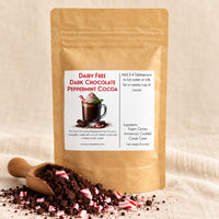 Thumbnail for A kraft paper stand-up pouch labeled 'Dairy Free Dark Chocolate Peppermint Cocoa' stands on a beige fabric surface with cocoa powder and candy cane pieces in a wooden scoop in front. The package has a white front label with red and black text and a photo of a hot chocolate cup.