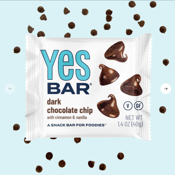 A Dark Chocolate Chip YES Bar® – Like a Cookie, But Better (and Guilt-Free!) is displayed in its wrapper with chocolate chips around it on a light blue background. The packaging highlights that its gluten-free, vegan, plant-based, and contains cinnamon and vanilla.