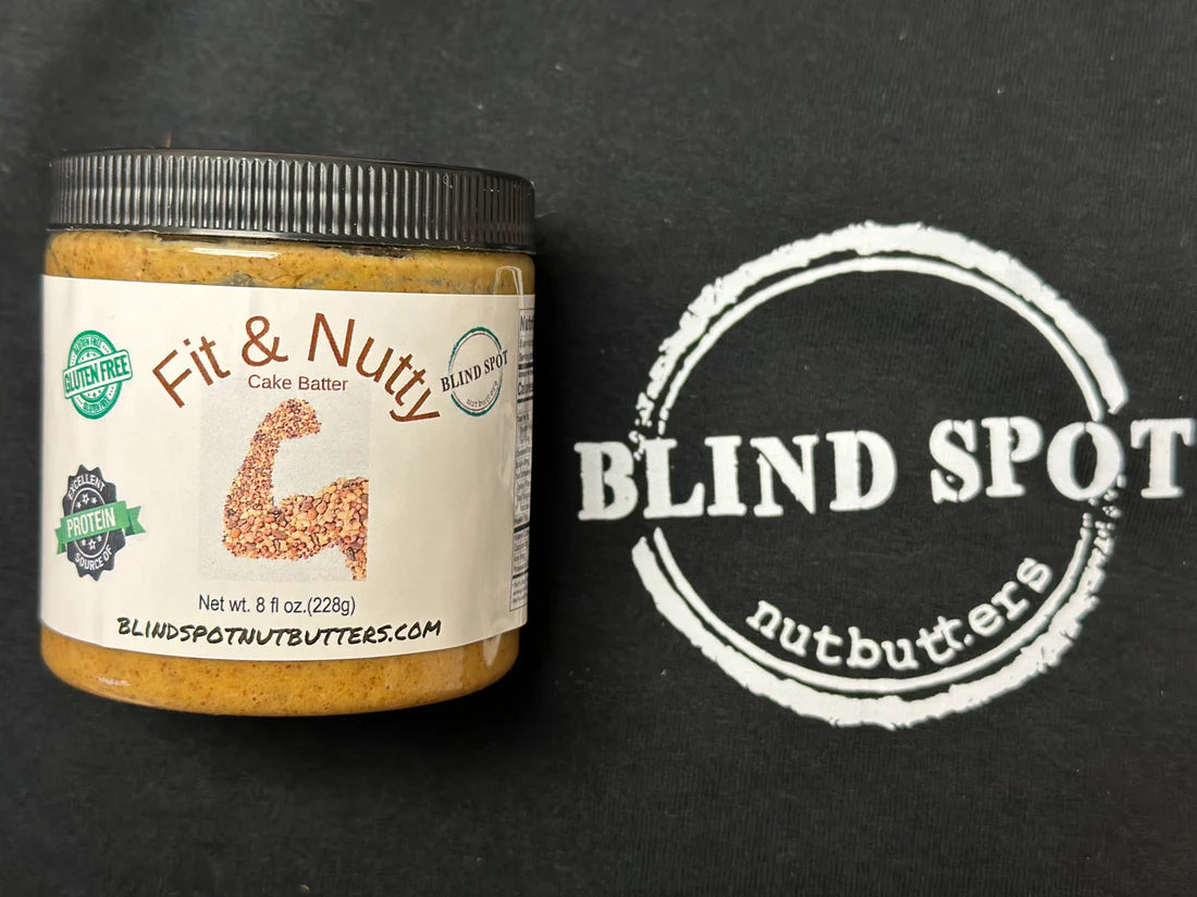 A jar of Fit & Nutty – 🎂 Vanilla Cake Batter Nut Butter – Sweet Gains & Birthday Vibes sits on a black surface with the Blind Spot Nutbutters logo and a flexed arm graphic made of sprinkled cake batter, emphasizing its high-protein content.