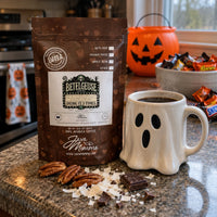 Thumbnail for A bag of Betelgeuse Java Momma coffee stands on a kitchen countertop beside a white ghost-shaped mug filled with black coffee. The scene includes Halloween-themed props like an orange jack-o'-lantern bucket, candy in a bowl, and scattered pecans and chocolate pieces.