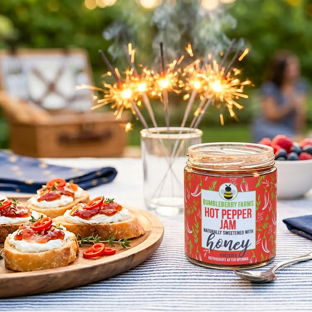 A jar of Rumbleberry Farms Hot Pepper Jam sits on a table outdoors beside bruschetta on a wooden board, with sparklers burning in the background.
