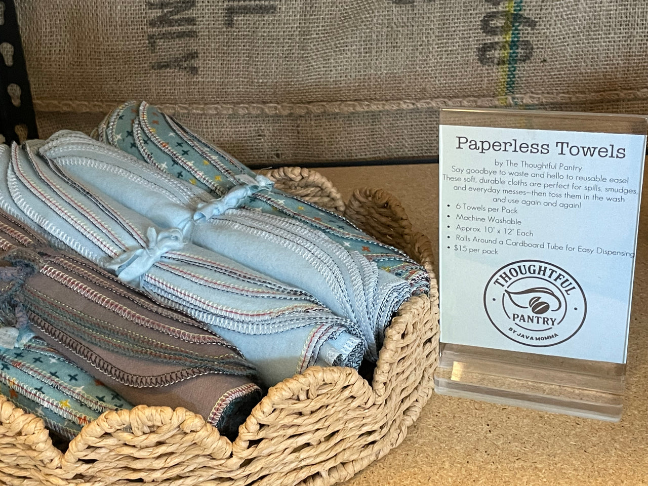 A woven basket holds folded, patterned Paperless Towels 🧻 – 6-Pack Reusable Cotton Flannel Towels by The Thoughtful Pantry with a sign noting their washable, eco-friendly, and affordable kitchen use.
