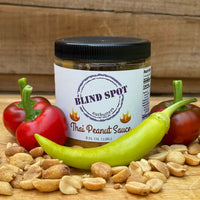 Thumbnail for Jar of Blind Spot Thai Peanut Sauce surrounded by peanuts and peppers on a wooden surface