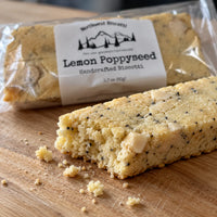 Thumbnail for A handcrafted biscotti bar with visible poppyseed and pale cream pieces, shown in the foreground on a wooden surface with crumbs scattered. Behind it is a clear plastic package labeled Northwest Biscotti Lemon Poppyseed Handcrafted Biscotti.