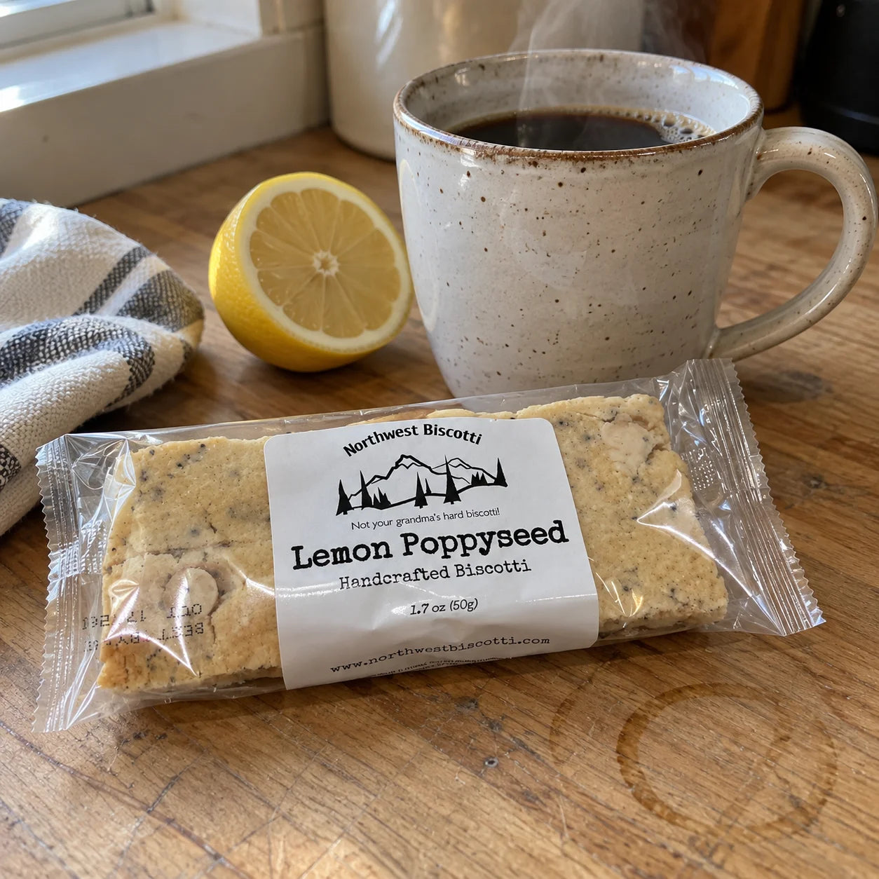 A packaged bar of Northwest Biscotti Lemon Poppyseed handcrafted biscotti sits on a wooden table in front of a mug of coffee, with a lemon half and a striped cloth nearby.