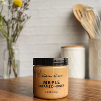 Thumbnail for A jar of Nate’s Nectar Maple 🍁 Creamed Honey – Cozy, Sweet & Spreadably Smooth rests on a wooden countertop, highlighting its rich maple flavor amid softly blurred kitchen utensils and a flower-filled glass vase in the background.