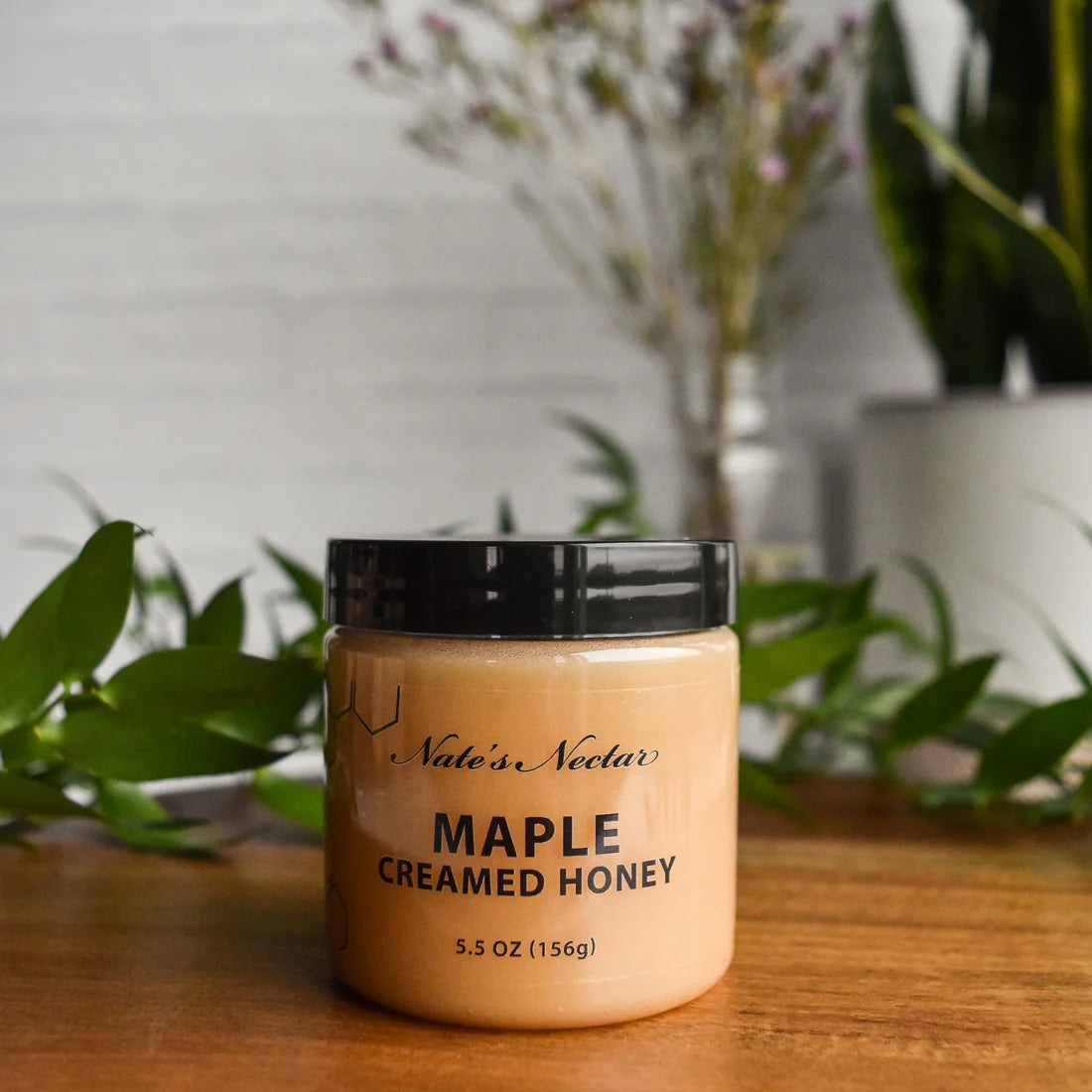 A 5.5 oz (156g) jar of Nate’s Nectar Maple 🍁 Creamed Honey – Cozy, Sweet & Spreadably Smooth sits on a wooden surface with green leaves in front. Its sleek black lid seals in raw, creamy honey—perfect for spreading or sweetening your favorite treats.