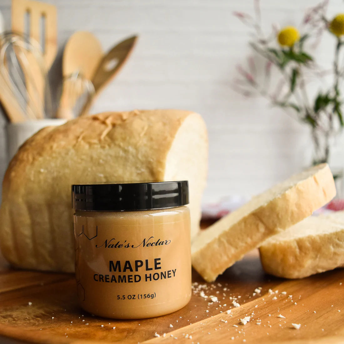 A jar of Nate’s Nectar Maple 🍁 Creamed Honey – Cozy, Sweet & Spreadably Smooth rests on wood before sliced bread, featuring this rich maple honey spread with softly blurred kitchen utensils and flowers in the background.