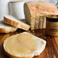 Thumbnail for A slice of bread with Nate’s Nectar Maple 🍁 Creamed Honey rests on a wooden board, while a loaf of homemade bread and the jar of this cozy, sweet & spreadably smooth treat are in the background.