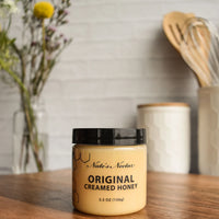 Thumbnail for A jar of Nate’s Nectar Original Creamed Honey 🐝 – Pure, Spreadable Bliss in a Jar rests on a wooden surface, kitchen utensils and flowers behind it. A black lid tops its honeycomb label, showcasing the smooth honey inside.