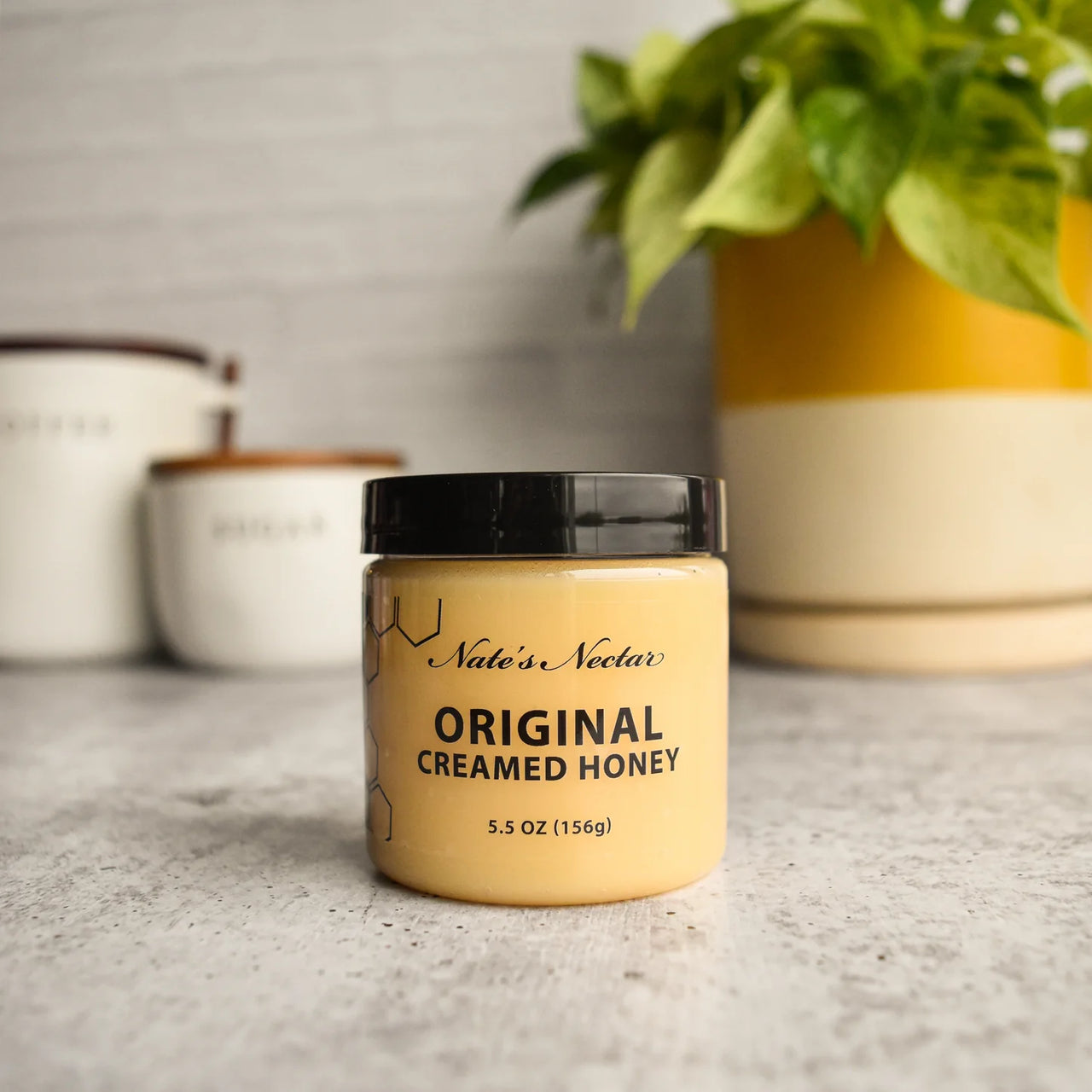 A jar of Nate’s Nectar Original Creamed Honey 🐝 – Pure, Spreadable Bliss in a Jar rests on a gray countertop, with two white canisters and a yellow-and-white pot holding a leafy green plant enhancing its smooth appeal.