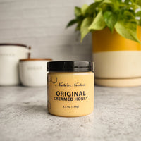 Thumbnail for A jar of Nate’s Nectar Original Creamed Honey 🐝 – Pure, Spreadable Bliss in a Jar rests on a gray countertop, with two white canisters and a yellow-and-white pot holding a leafy green plant enhancing its smooth appeal.
