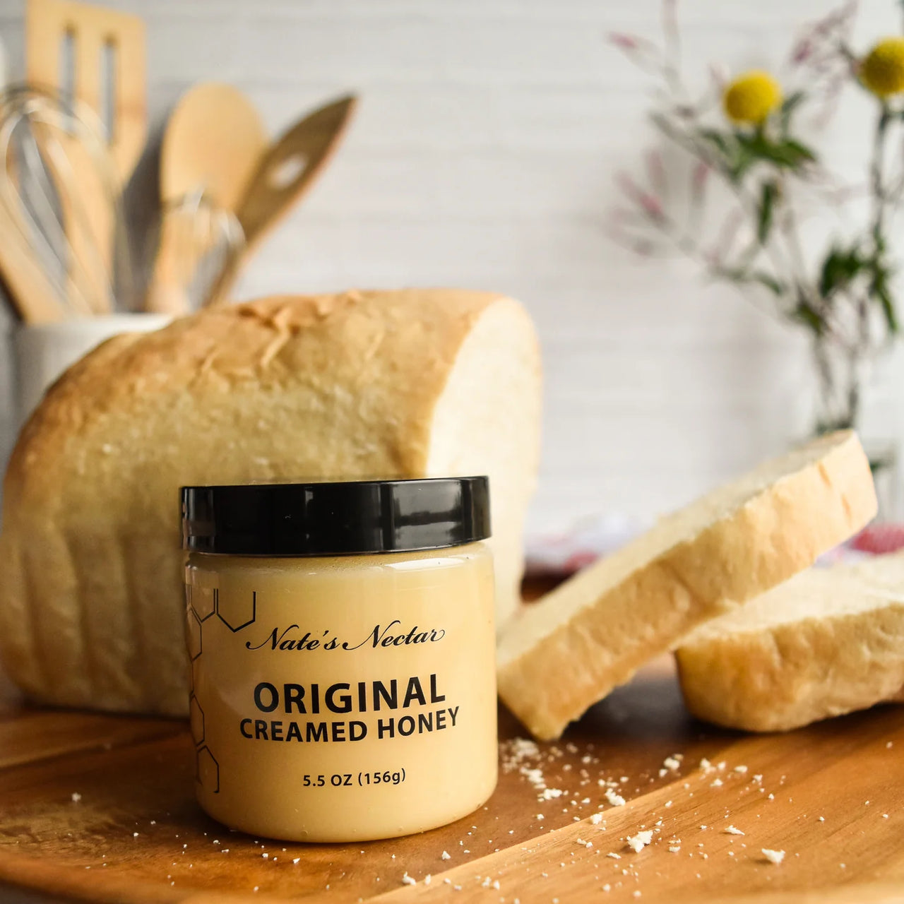 A jar of Nate’s Nectar Original Creamed Honey 🐝 – Pure, Spreadable Bliss in a Jar rests on a wooden board next to sliced bread, ready to elevate your breakfast. Kitchen utensils and a flower vase are softly blurred in the background.