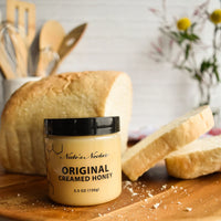 Thumbnail for A jar of Nate’s Nectar Original Creamed Honey 🐝 – Pure, Spreadable Bliss in a Jar rests on a wooden board next to sliced bread, ready to elevate your breakfast. Kitchen utensils and a flower vase are softly blurred in the background.