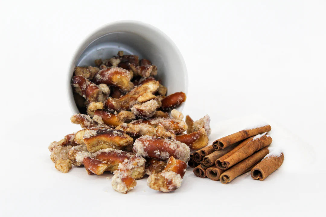 A white cup spills Sugar + Spice Pretzel Bites – Sweet, Salty, and Straight-Up Craveable 🍬🔥 onto a white surface next to cinnamon sticks; these small-batch bites are coated in delicious cinnamon sugar.