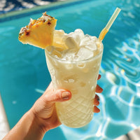 Thumbnail for A hand holds a glass of iced, creamy pineapple drink with Coconut Skinny Syrup – A Tropical Dream in Every Drop 🥥🏝️✨, garnished with pineapple and a straw, by a sparkling pool. Enjoy this keto-friendly, coconut-flavored treat!.
