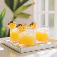 Thumbnail for Three glasses of yellow juice with ice, each topped with a pineapple wedge, sit on a white tray in the sun—sweetened by Pineapple Upside Down Cake Skinny Syrup – Dessert Meets Paradise 🍍🍰✨—with blurred green plants in the background.