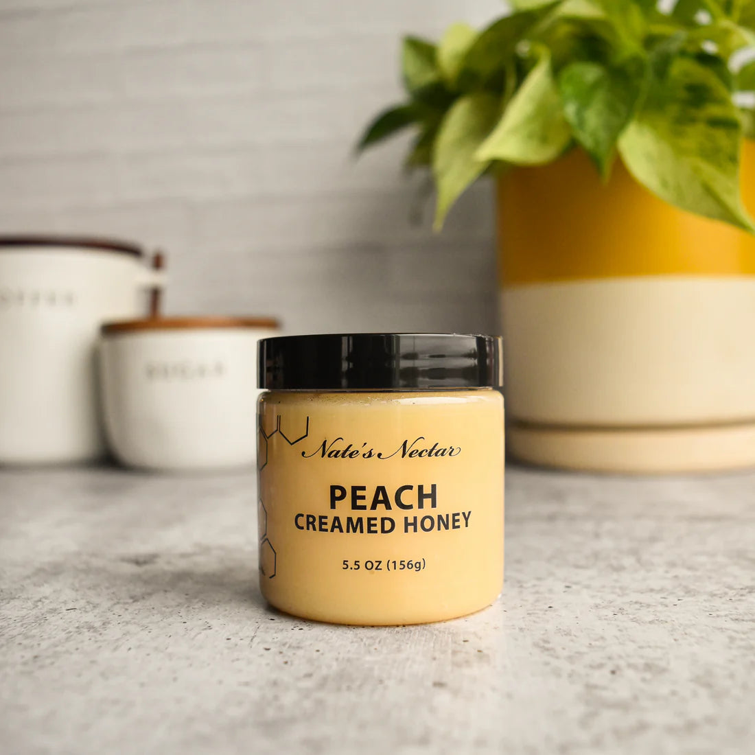 A jar of Nate’s Nectar Peach 🍑 Creamed Honey – Juicy, Sweet & Perfectly Spreadable rests on a gray countertop, with a green plant and two labeled canisters blurred in the background.