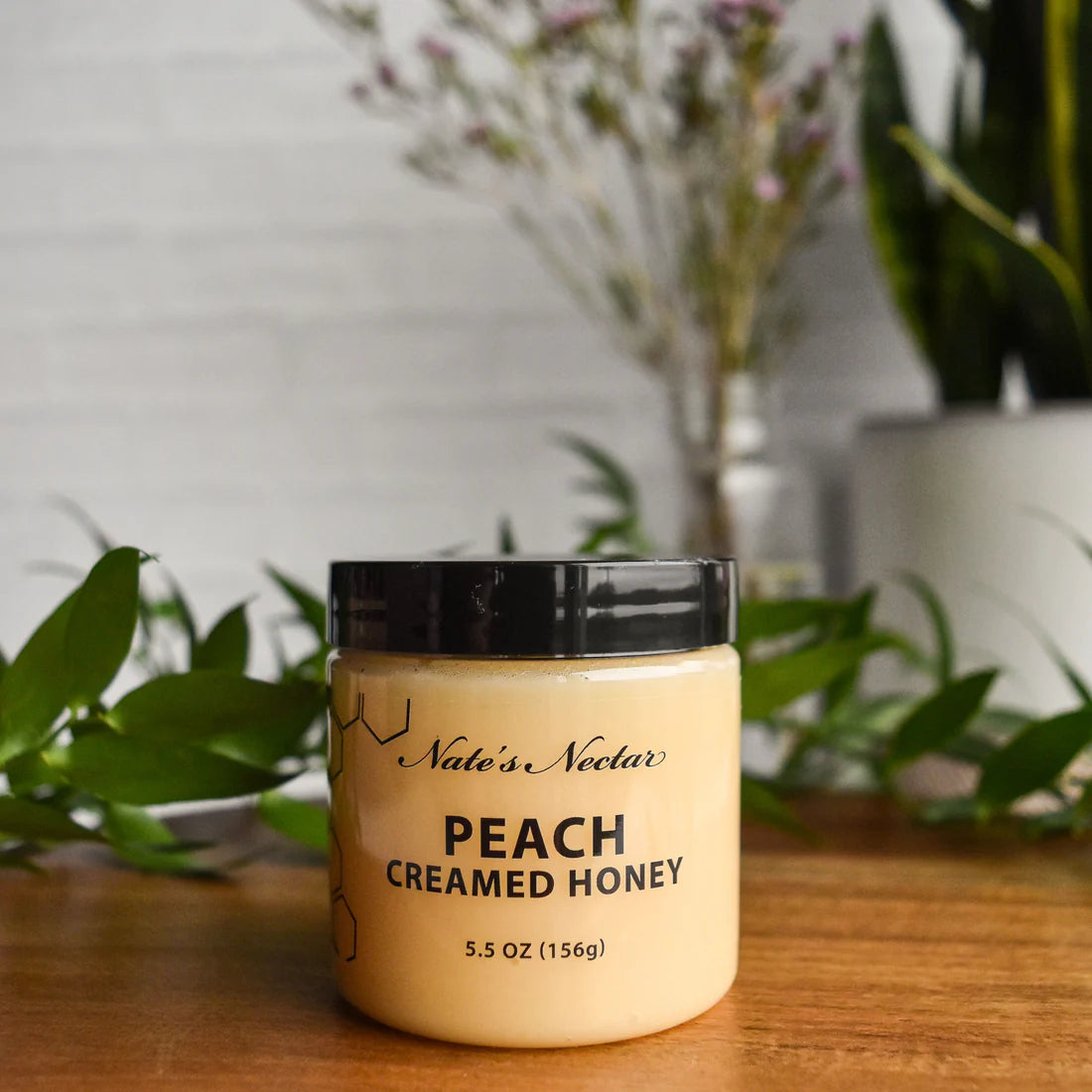 A jar of Nate’s Nectar Peach 🍑 Creamed Honey – Juicy, Sweet & Perfectly Spreadable sits on a wooden surface among green plants, with a soft-focus background of flowers and a white brick wall.