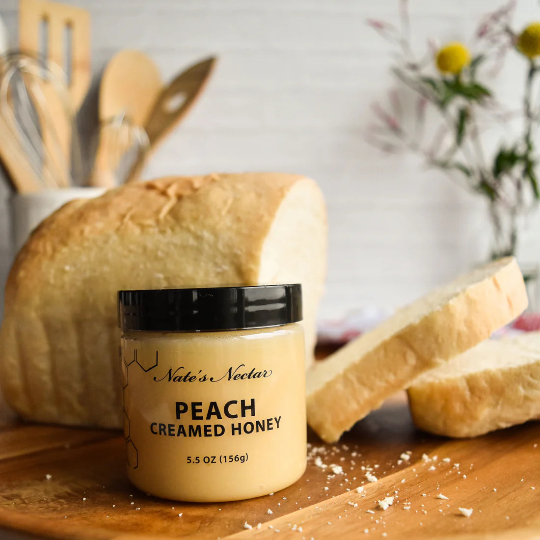 A jar of Nate’s Nectar Peach 🍑 Creamed Honey – Juicy, Sweet & Perfectly Spreadable sits in front of sliced bread on a kitchen counter, surrounded by wooden utensils and flowers.
