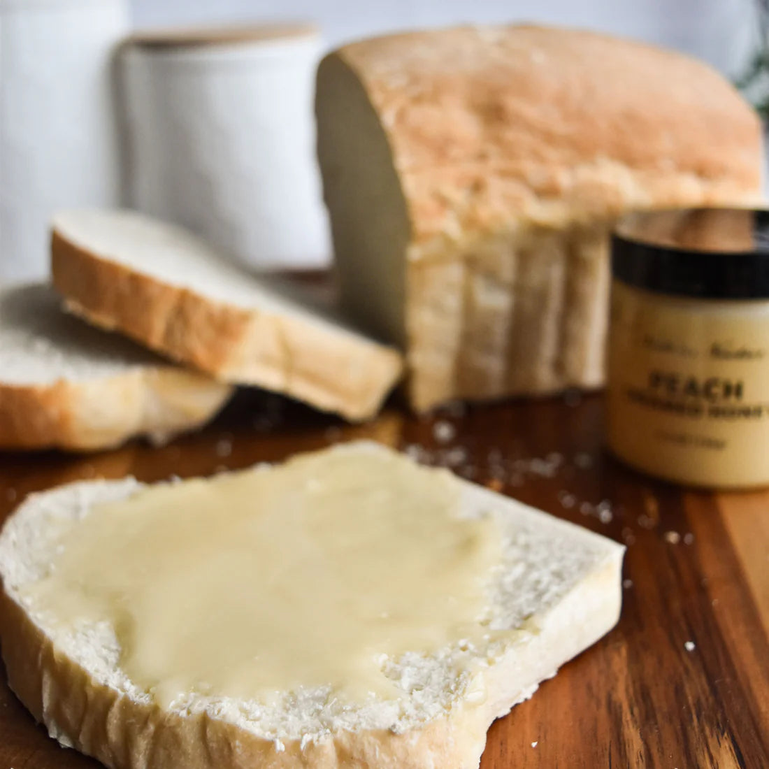 A slice of white bread with creamy butter sits on a wooden board alongside more slices, a loaf, and a jar of Nate’s Nectar Peach 🍑 Creamed Honey – Juicy, Sweet & Perfectly Spreadable in the background.