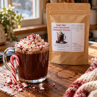 Thumbnail for A kraft paper pouch of Dairy Free Dark Chocolate Peppermint Cocoa stands next to a glass mug filled with the prepared cocoa, topped with whipped cream, candy canes, and sprinkles on a wooden table.