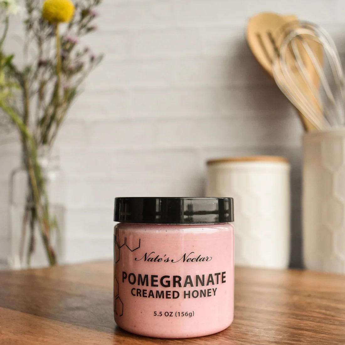 A jar of Nate’s Nectar Pomegranate Creamed Honey – Bold, Sweet & Irresistibly Smooth rests on a wooden countertop, with blurred kitchen utensils and a vase of flowers in the background.