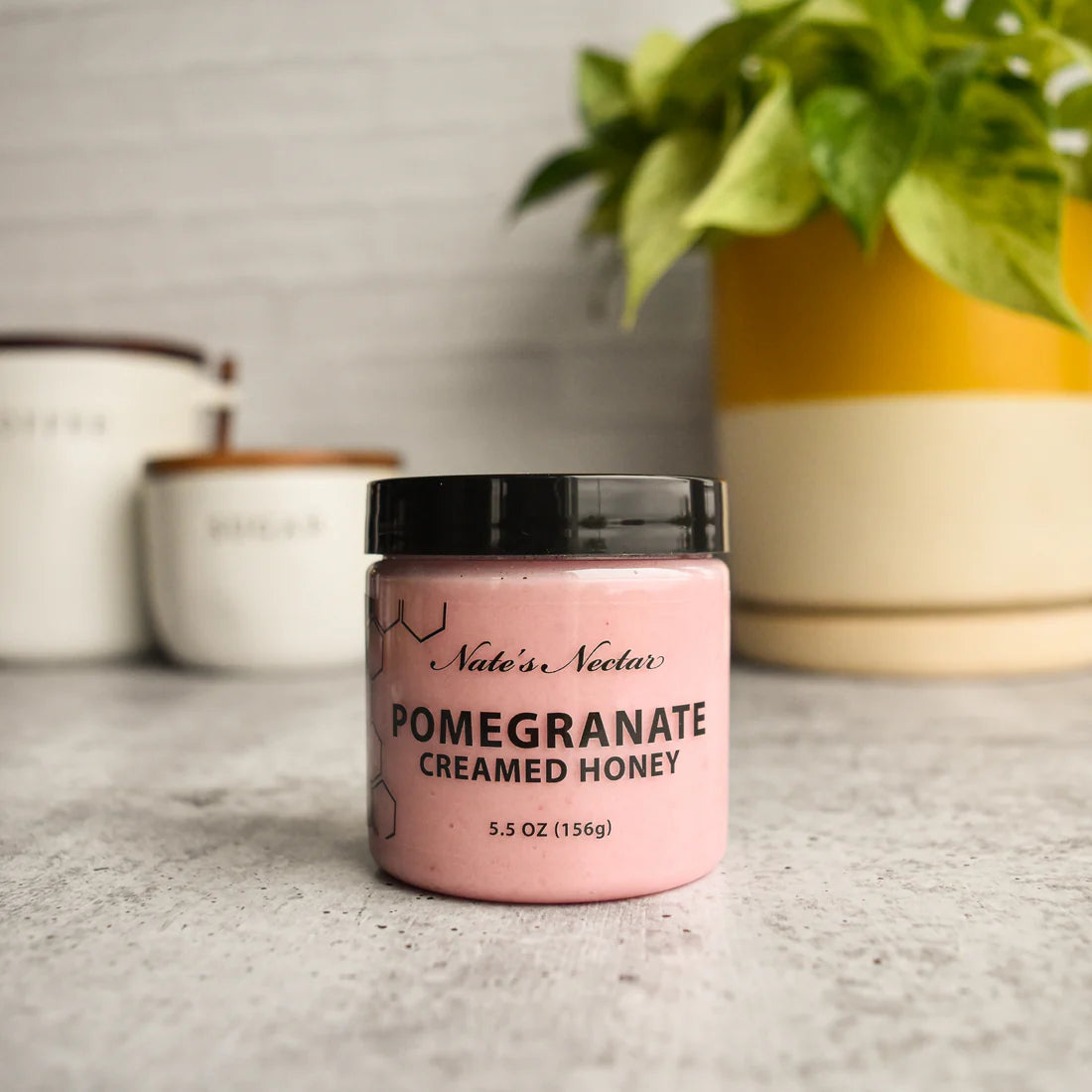 A jar of Nate’s Nectar Pomegranate Creamed Honey – Bold, Sweet & Irresistibly Smooth sits on a gray countertop, with houseplants and labeled kitchen canisters blurred in the background.
