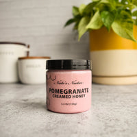 Thumbnail for A jar of Nate’s Nectar Pomegranate Creamed Honey – Bold, Sweet & Irresistibly Smooth sits on a gray countertop, with houseplants and labeled kitchen canisters blurred in the background.