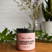 Thumbnail for A jar of Nate’s Nectar Pomegranate Creamed Honey – Bold, Sweet & Irresistibly Smooth, sits on a wooden surface, surrounded by green leaves in the foreground and blurred plants and flowers in the background.