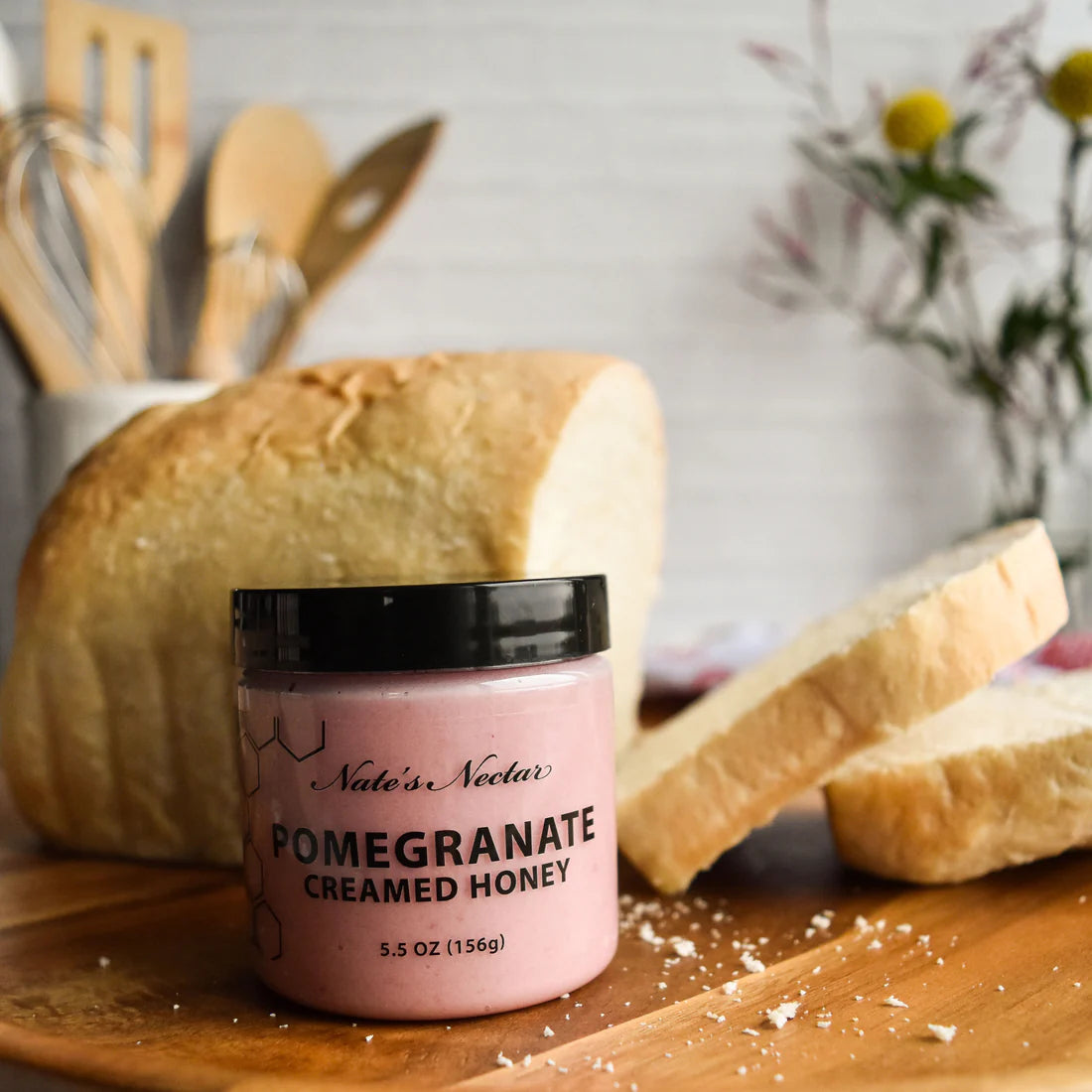 A jar of Nate’s Nectar Pomegranate Creamed Honey – Bold, Sweet & Irresistibly Smooth sits on a wooden surface beside sliced bread, with blurred kitchen utensils and flowers in the background, showcasing this unique fruity honey spread.