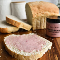 Thumbnail for A slice of white bread topped with Nate’s Nectar Pomegranate Creamed Honey sits on a wooden surface, with extra slices, a loaf of bread, and the jar labeled “Nate’s Nectar Pomegranate Creamed Honey – Bold, Sweet & Irresistibly Smooth” behind it.