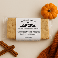 Thumbnail for A Pumpkin Spice Walnut Biscotti Double Pack (Soft-Bite, Spiced, & Seriously Snackable) is shown with a small pumpkin, two cinnamon sticks, and orange knit fabric on beige. The label highlights soft-baked perfection—not your grandma’s hard biscotti.