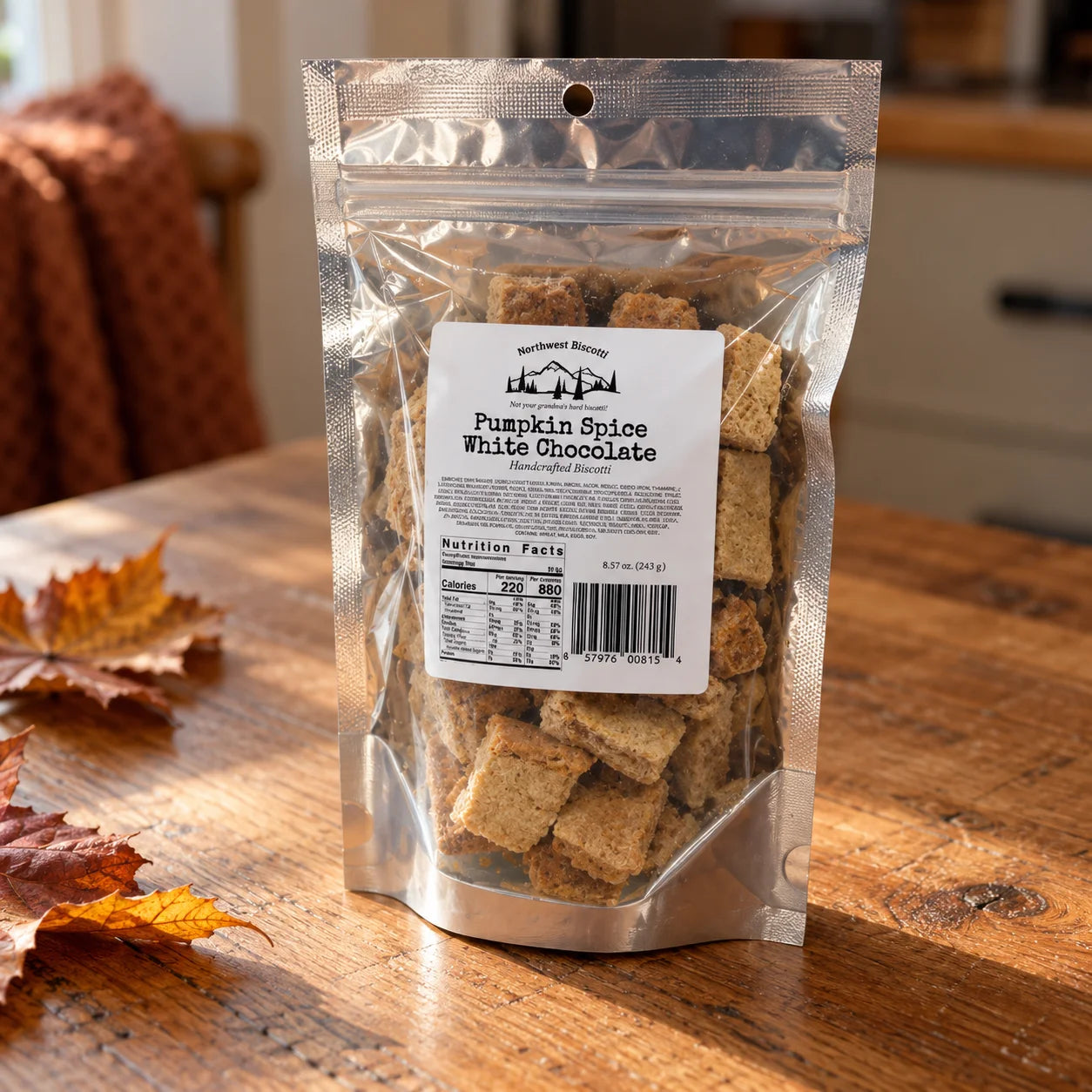 A stand-up resealable pouch of Northwest Biscotti Pumpkin Spice White Chocolate Handcrafted Biscuits sits on a wooden table with autumn leaves nearby. The clear bag shows the baked biscuits inside, with a white label displaying the brand and product name.