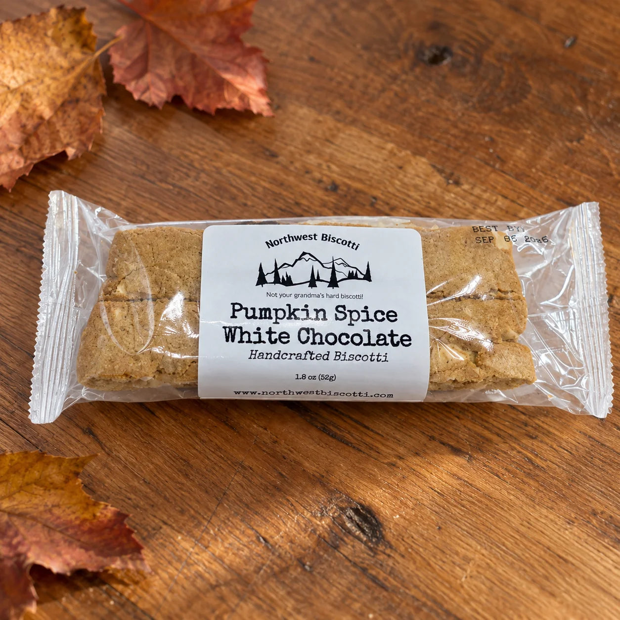 A packaged biscotti cookie on a wooden surface with autumn leaves. The clear wrapper shows two tan-brown biscotti bars and a white label reading Northwest Biscuits Pumpkin Spice White Chocolate Handcrafted Biscotti.