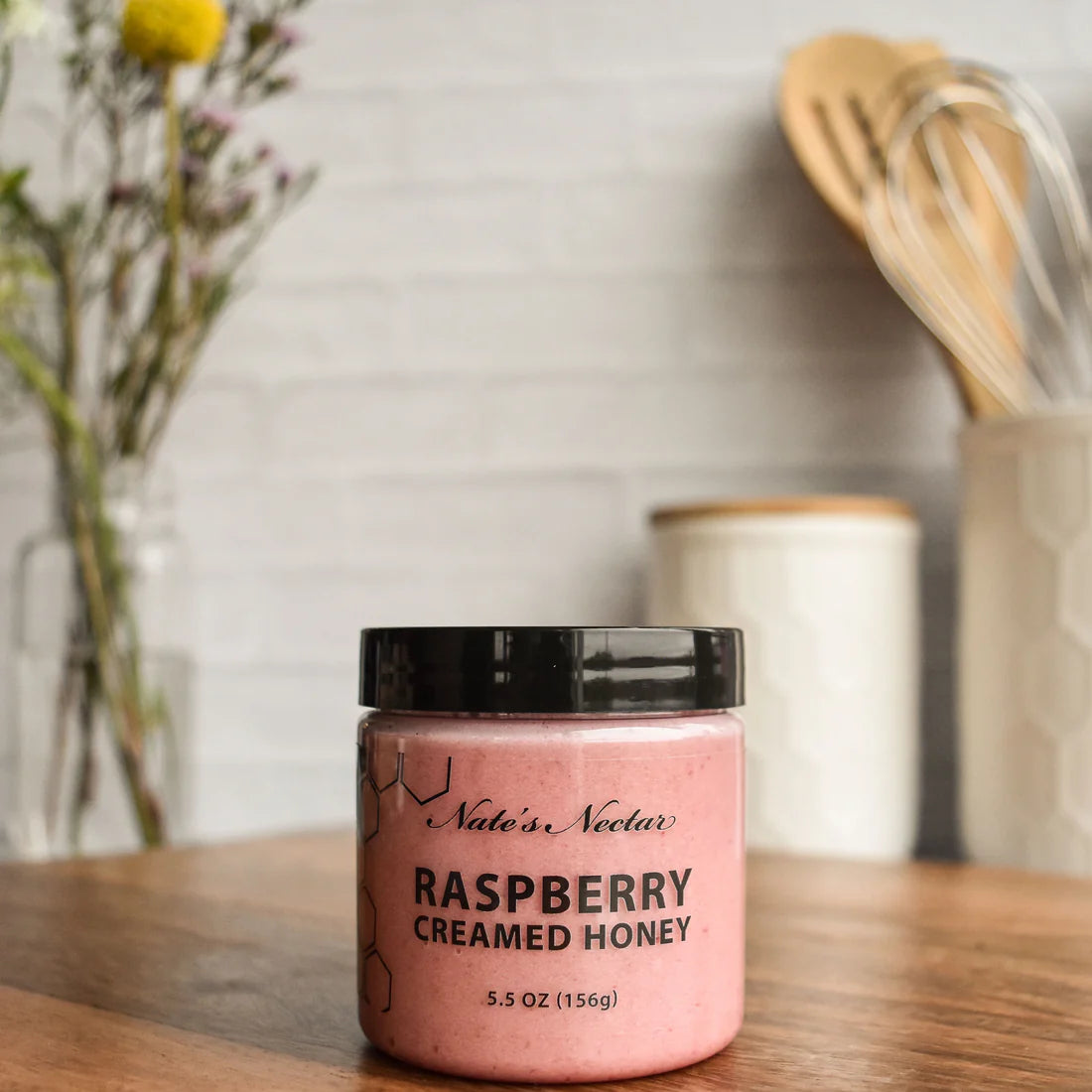 A jar of Nate’s Nectar Raspberry Creamed Honey – Tangy, Sweet & Spreadably Smooth sits on a wooden countertop, surrounded by kitchen utensils and flowers. The pink honey is in a clear jar with a black lid and black label text.
