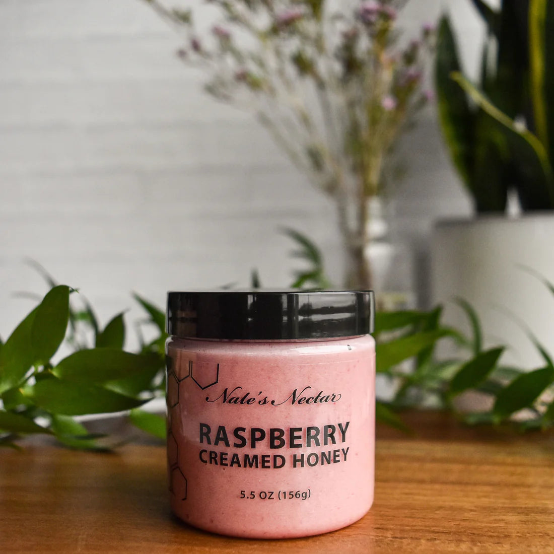 A jar of Nate’s Nectar Raspberry Creamed Honey – Tangy, Sweet & Spreadably Smooth rests on a wooden surface with green leaves and blurred plants behind it. Pink honey is visible in the clear jar with a black lid and label.