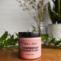 Thumbnail for A jar of Nate’s Nectar Raspberry Creamed Honey – Tangy, Sweet & Spreadably Smooth rests on a wooden surface with green leaves and blurred plants behind it. Pink honey is visible in the clear jar with a black lid and label.