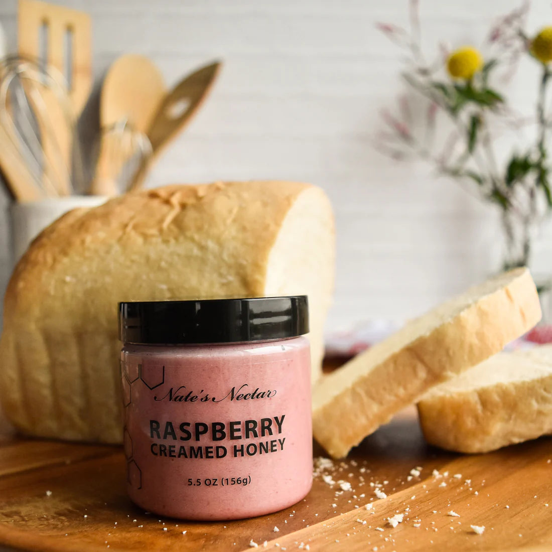 A jar of Nate’s Nectar Raspberry Creamed Honey – Tangy, Sweet & Spreadably Smooth rests on a wooden board before a loaf of sliced bread, with blurred kitchen utensils and flowers in the background.