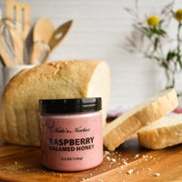 Thumbnail for A jar of Nate’s Nectar Raspberry Creamed Honey – Tangy, Sweet & Spreadably Smooth rests on a wooden board before a loaf of sliced bread, with blurred kitchen utensils and flowers in the background.