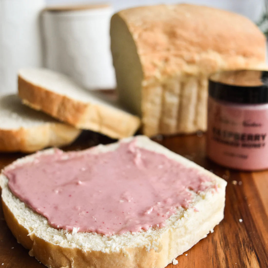A slice of white bread with Nate’s Nectar Raspberry Creamed Honey – Tangy, Sweet & Spreadably Smooth sits on a wooden surface, surrounded by more sliced bread, a loaf, and a jar of the honey in the background.