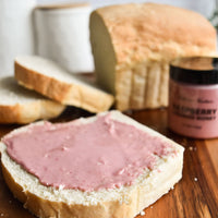 Thumbnail for A slice of white bread with Nate’s Nectar Raspberry Creamed Honey – Tangy, Sweet & Spreadably Smooth sits on a wooden surface, surrounded by more sliced bread, a loaf, and a jar of the honey in the background.