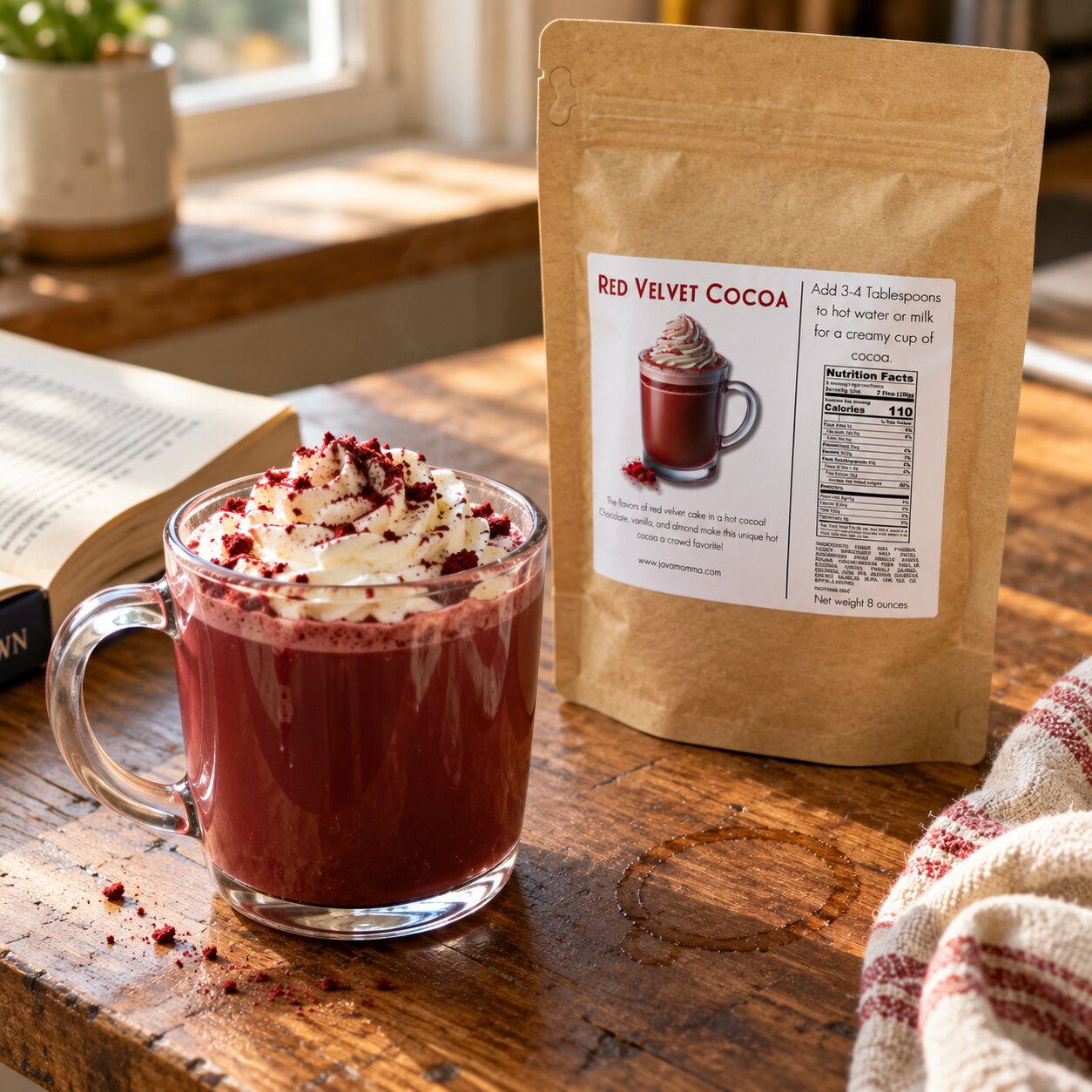 A kraft paper pouch labeled Red Velvet Cocoa stands on a wooden table beside a glass mug filled with a red velvet hot cocoa drink, topped with whipped cream and cocoa powder, with an open book and soft window light in the background.