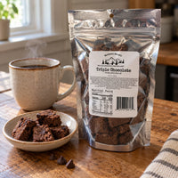 Thumbnail for A bag of Northwest Biscotti Triple Chocolate Brownies stands on a wooden table beside a ceramic mug of coffee and a small bowl of brownie pieces. The product packaging is a silver resealable pouch with a white label.