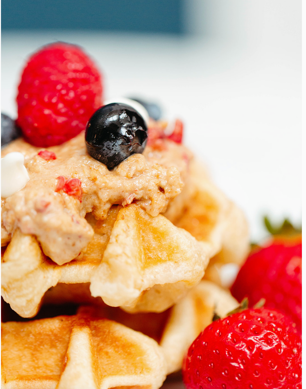 A close-up of waffles stacked with 🍓 Raspberry White Chocolate BnuttyPeanut Butter – Tart, Sweet, and Totally Luxe, topped with berries and cream, plus fresh strawberries and more berries on the side.
