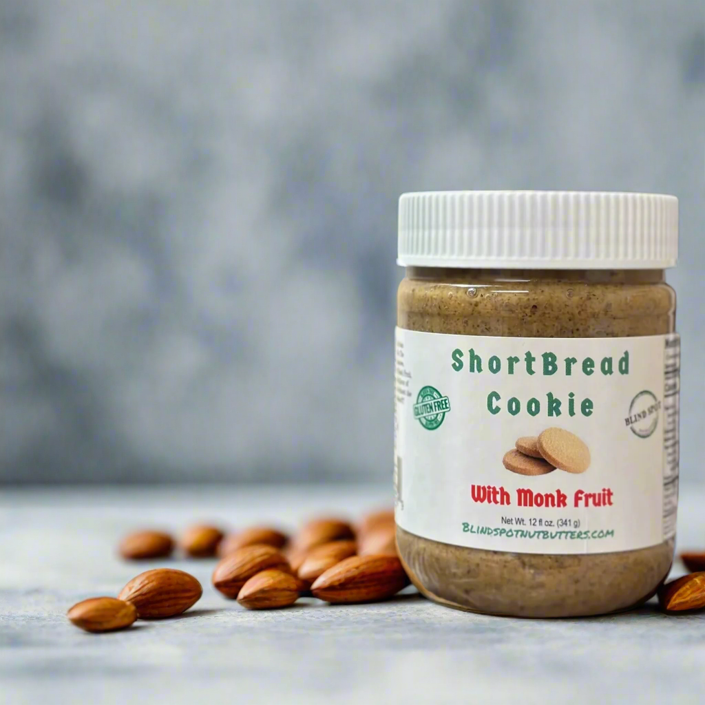 A jar of Shortbread Cookie 🫓 with Monk Fruit Nut Butter – Buttery, Sweet & Sugar-Free Bliss sits on a gray surface among whole almonds, set against a softly blurred, neutral background.