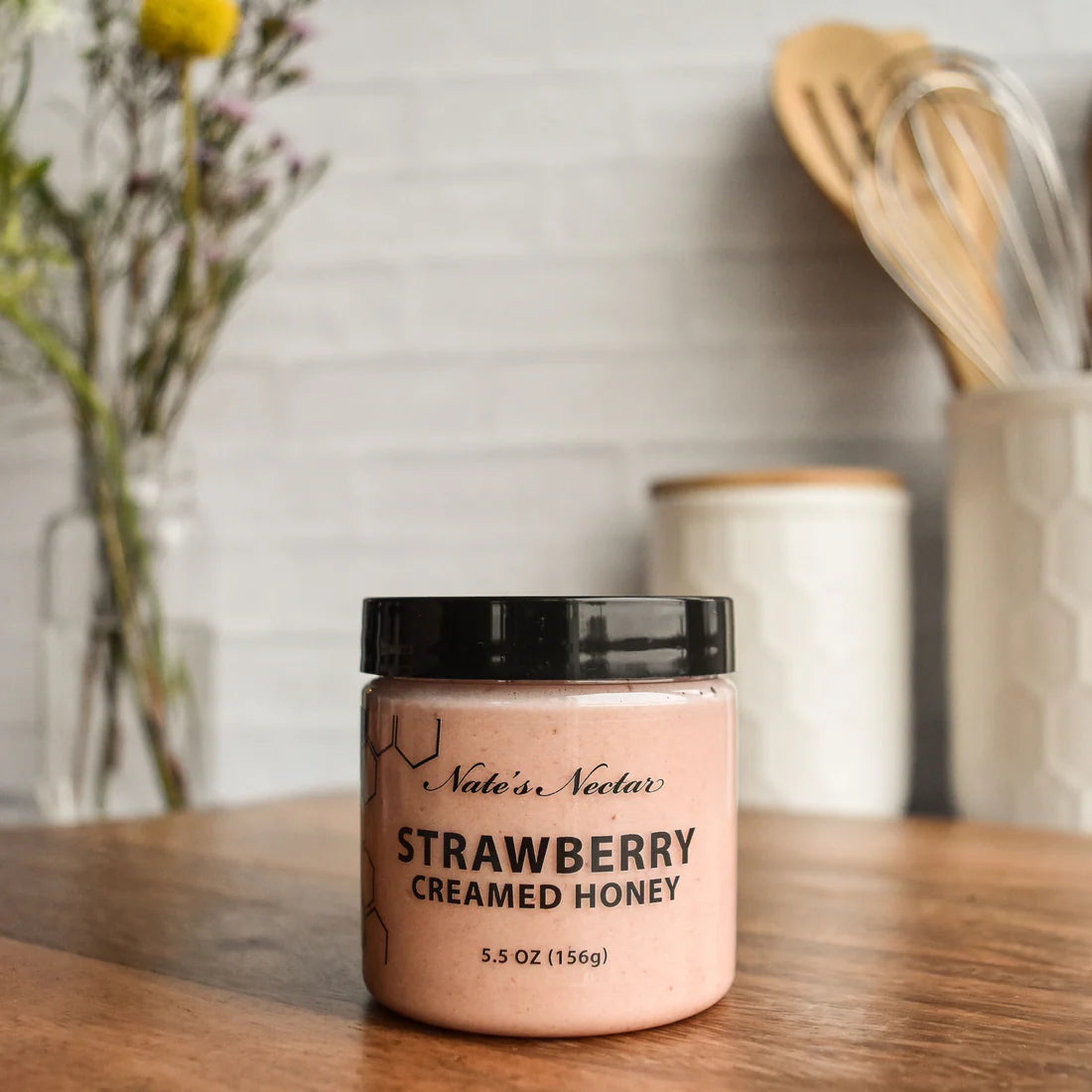 A jar of Nate’s Nectar Strawberry Creamed Honey 🍓 – Sweet, Fruity, and Ready to Spread rests on a wooden surface, while kitchen utensils, a white canister, and flowers create a softly blurred background.