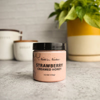 Thumbnail for A jar of Nate’s Nectar Strawberry Creamed Honey 🍓 – Sweet, Fruity, and Ready to Spread – rests on a gray countertop beside a plant in a yellow and white pot with two ceramic canisters blurred in the background.