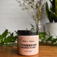 Thumbnail for A jar of Nate’s Nectar Strawberry Creamed Honey 🍓 – Sweet, Fruity, and Ready to Spread sits on a wooden table among green leaves, with plants in the background—perfect as a berry spread or for enjoying raw honey flavor.