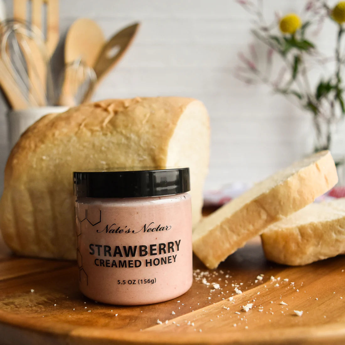 A jar of Nate’s Nectar Strawberry Creamed Honey 🍓 – Sweet, Fruity, and Ready to Spread – sits on a wooden board beside white bread slices, with kitchen utensils and flowers softly blurred in the background.